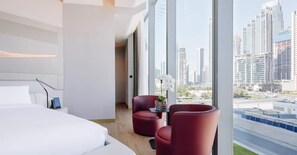 A modern hotel room with a large bed, two red chairs, a small table with a vase, and a city view through large windows. - ME by Melia Dubai (Dubai)