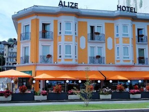 A hotel with a yellow facade, balconies with flower boxes, and outdoor seating with orange umbrellas. - Hotel Alize Evian (Evian-les-Bains)