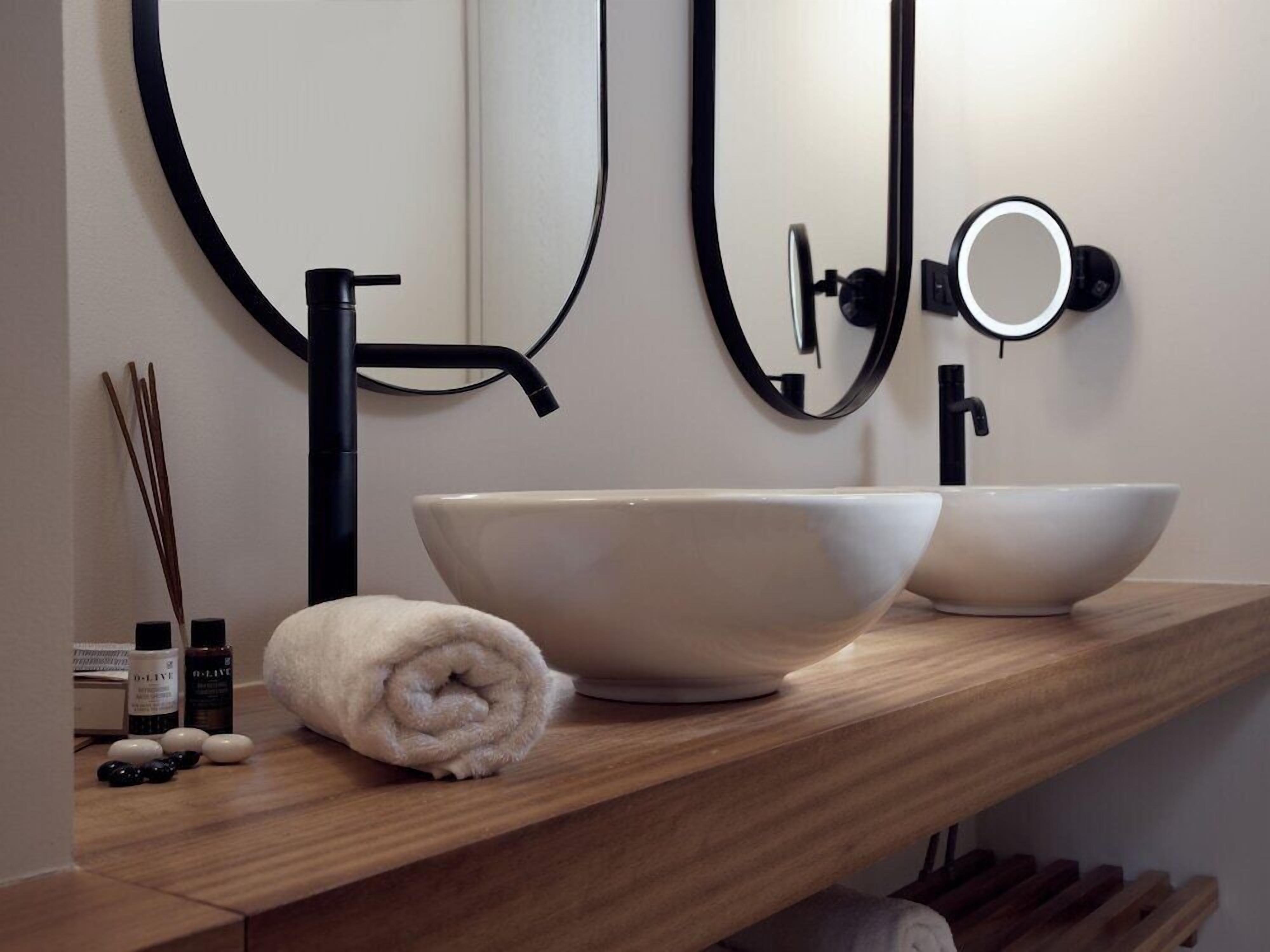 A modern bathroom with a wooden vanity, two white vessel sinks, a black faucet, and a towel rolled up on the counter.