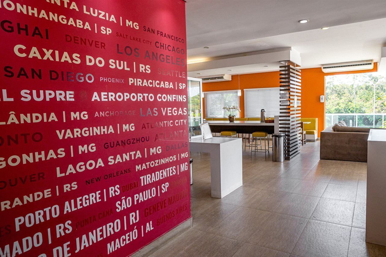Un vestíbulo de hotel con una amplia pared roja que muestra nombres de ciudades, un área de descanso con mesas y sillas, y vistas del exterior a través de grandes ventanales.