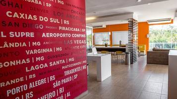 Un vestíbulo de hotel con una amplia pared roja que muestra nombres de ciudades, un área de descanso con mesas y sillas, y vistas del exterior a través de grandes ventanales.