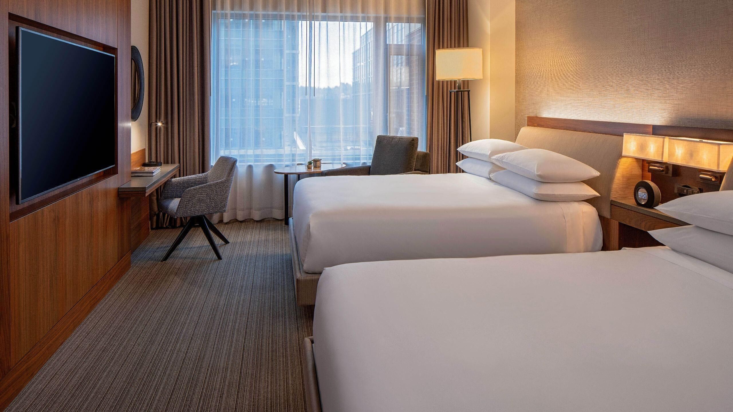 Quarto de hotel com duas camas, uma mesa, uma cadeira, uma televisão e janela com cortinas.