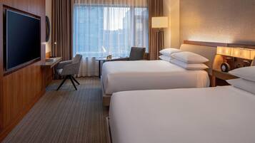 Quarto de hotel com duas camas, uma mesa, uma cadeira, uma televisão e janela com cortinas.