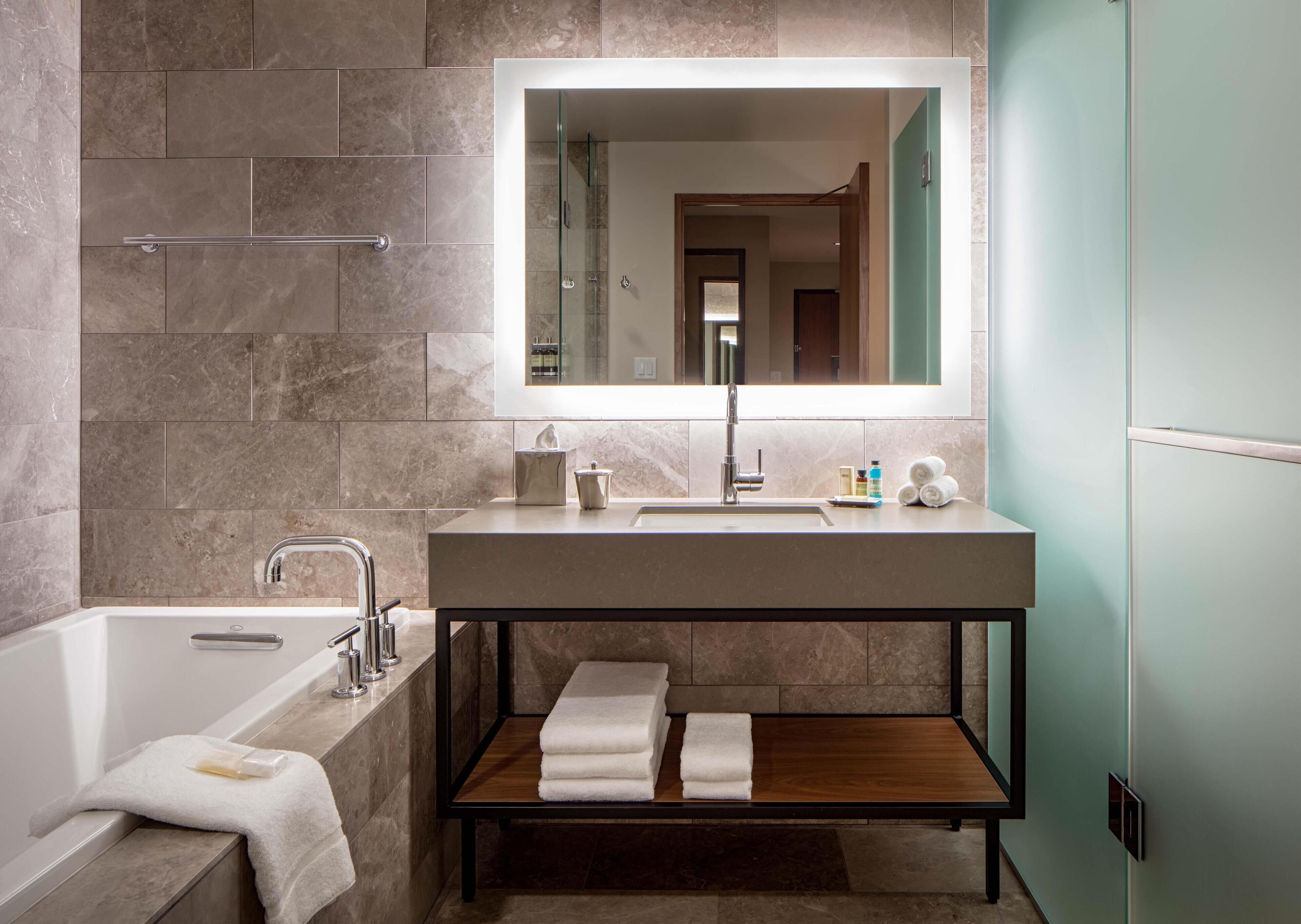 A modern bathroom with a bathtub, a large mirror, a vanity with a sink, and a shelf with towels.