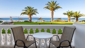 A terrace with a white table and two chairs, overlooking a beach with palm trees and umbrellas. - Marina Bay Hotel Aparthotel (Kefalonia)