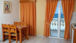 Una habitación con una mesa y sillas de madera, cortinas anaranjadas, vista al mar a través de un ventanal y una pequeña cocina.