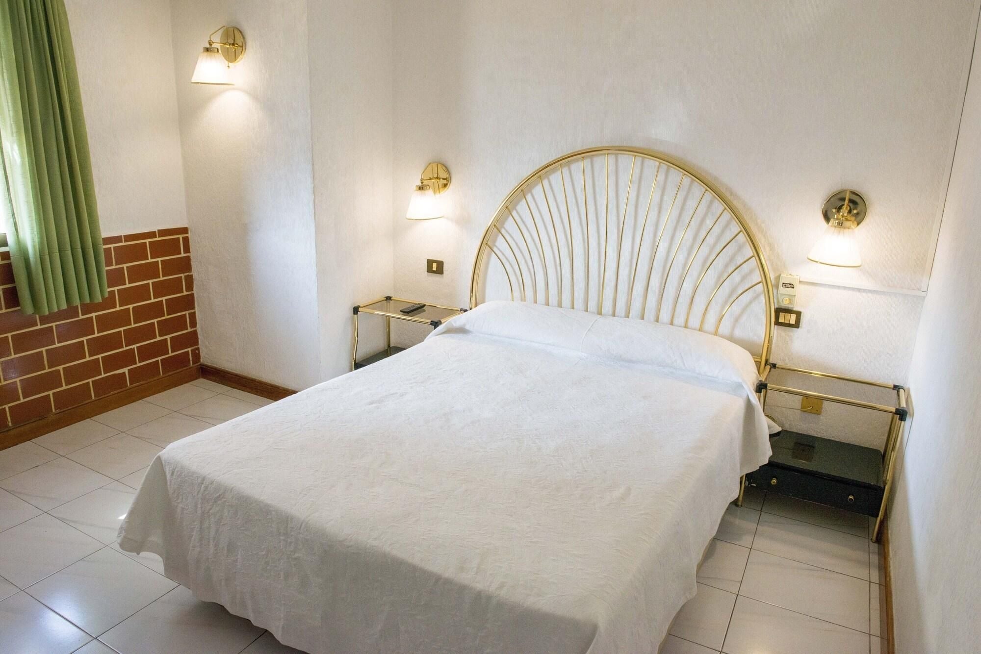 Ein ordentlich bezogenes Bett mit weißer Bettdecke in einem Hotelzimmer. Es befinden sich zwei Nachttische mit Lampen, eine Ziegelwand und ein grüner Vorhang im Raum.