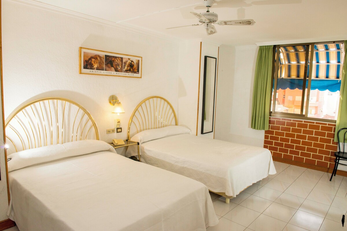 Ein Hotelzimmer mit zwei Einzelbetten, einem Deckenventilator, einem Fenster mit gestreifter Gardine und einem gerahmten Bild an der Wand.