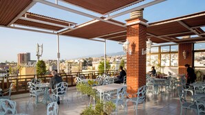 Een terras met metalen stoelen en tafels, een bakstenen pilaar en uitzicht op de stad.