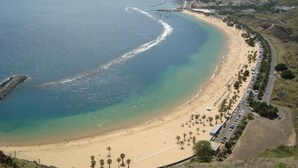 Una playa costera con una carretera que recorre la orilla, palmeras y un paisaje montañoso al fondo.