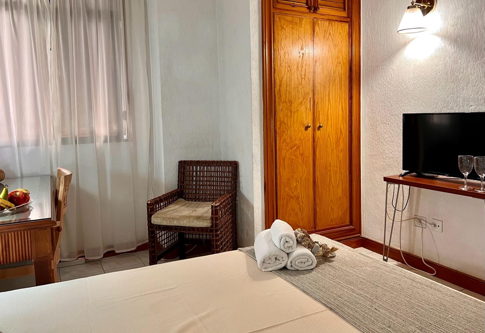 A hotel room with a bed, a wicker chair, a wooden wardrobe, a television, and a small table with fruit and towels.