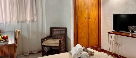 Habitación de hotel con cama, una silla de mimbre, un armario de madera, un televisor y una mesita con fruta y toallas.