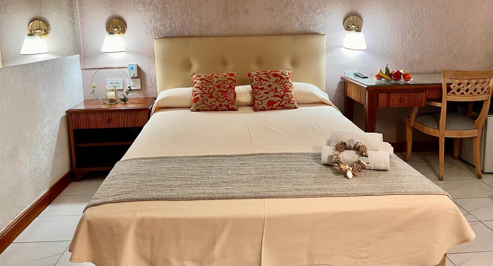 Quarto de hotel com uma cama grande, duas almofadas, mesa de cabeceira com abajur, uma cadeira e uma pequena mesa com frutas.
