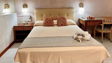 Quarto de hotel com uma cama grande, duas almofadas, mesa de cabeceira com abajur, uma cadeira e uma pequena mesa com frutas.