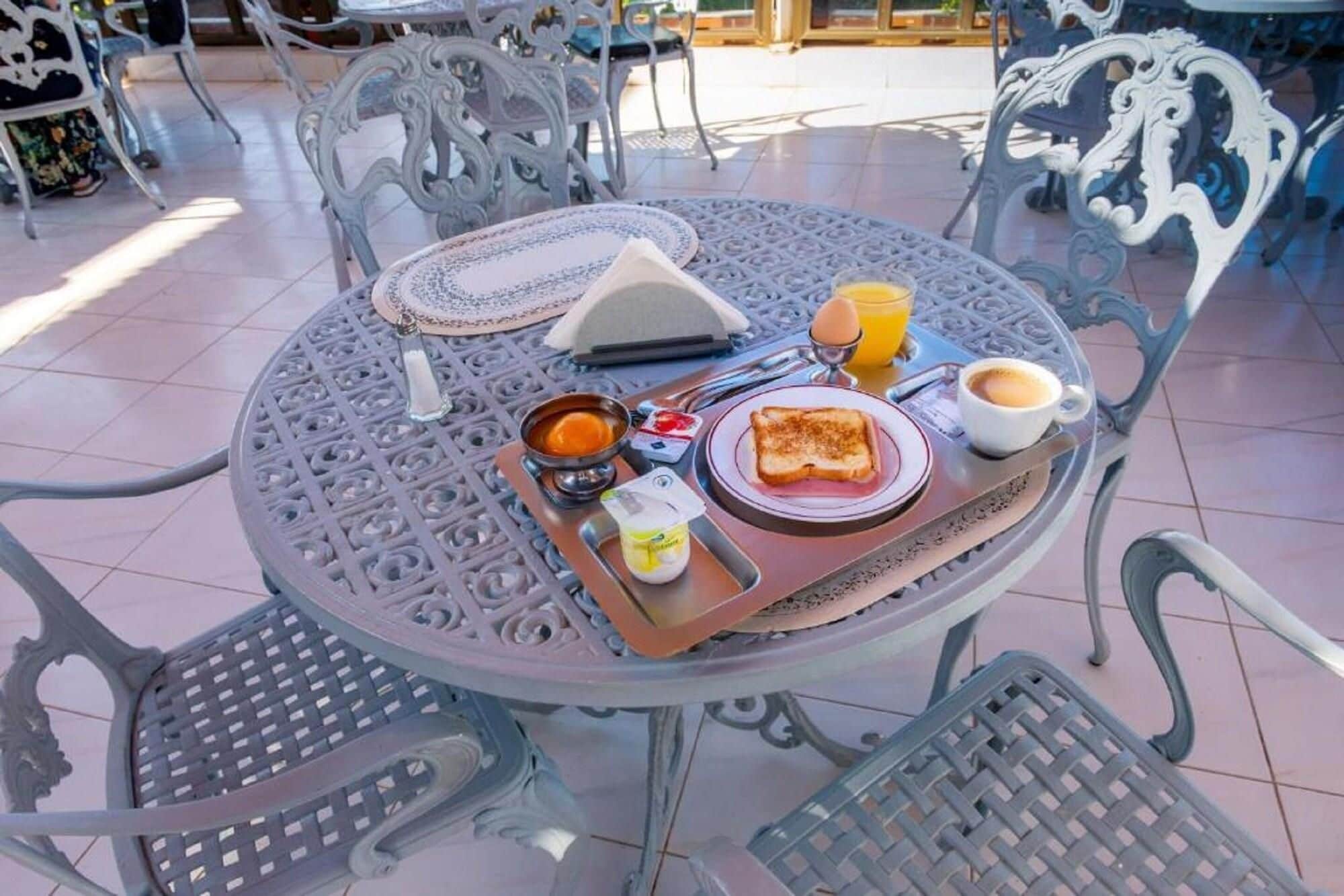 Una mesa redonda de metal con una bandeja de alimentos para el desayuno, como tostadas, huevos y jugo, rodeada de sillas de metal decorativas.