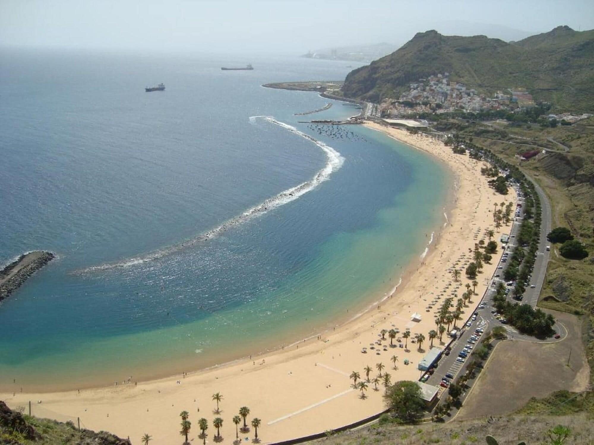A coastal beach with a road running parallel to the shore, palm trees, and a hilly landscape in the background.