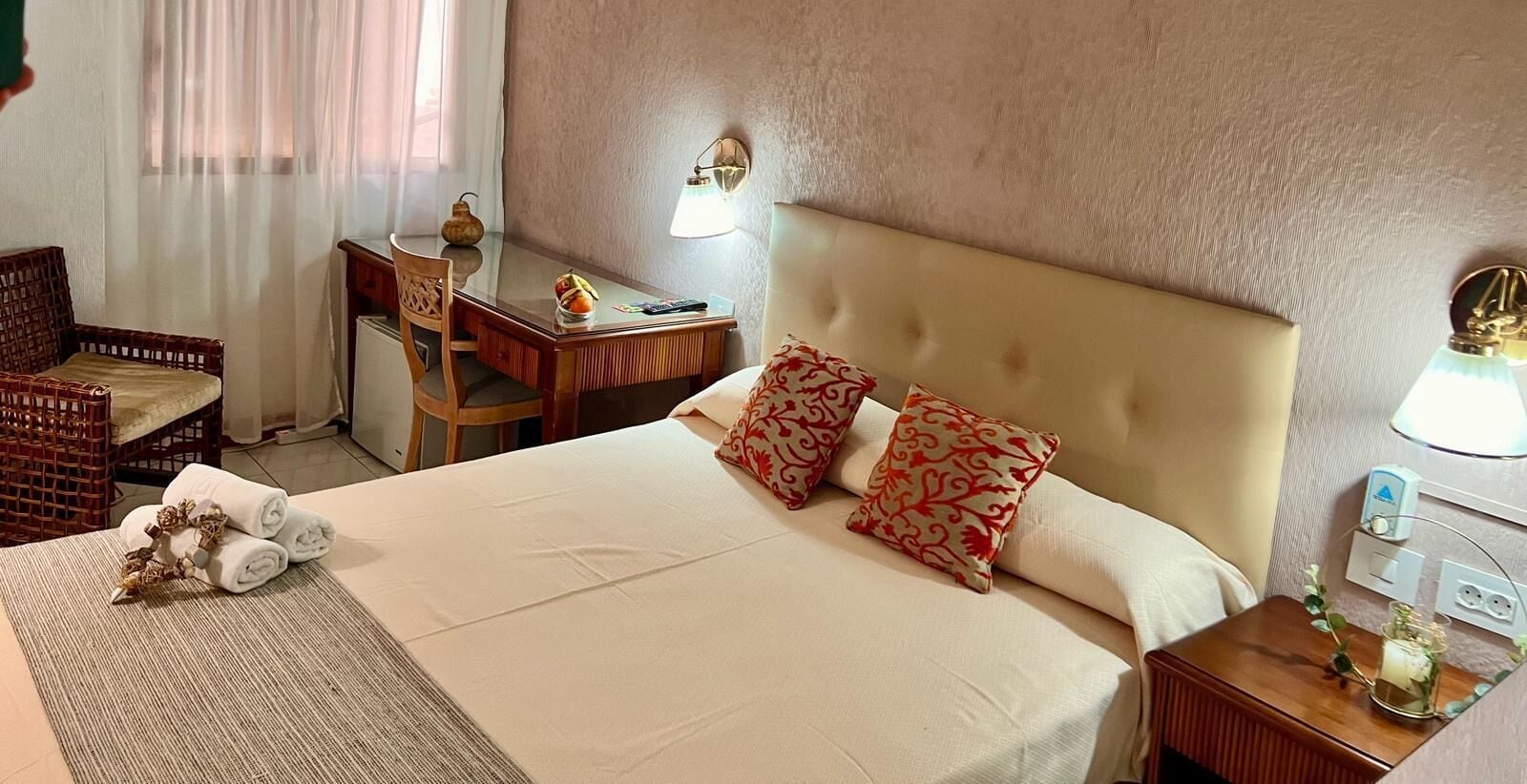 Habitación de hotel con una cama grande, dos almohadas, un escritorio con lámpara, una silla y una mesita con un jarrón.