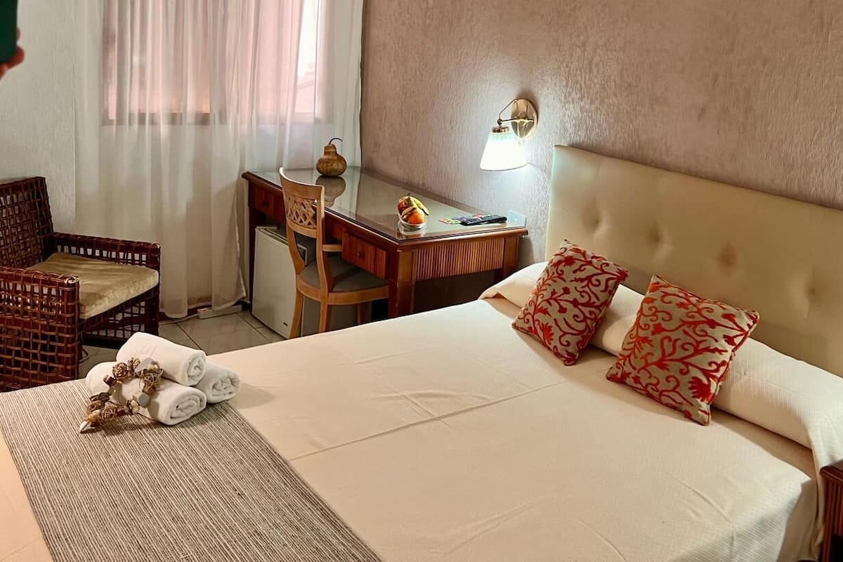 Habitación de hotel con una cama grande, dos almohadas, un escritorio con lámpara, una silla y una mesita con un jarrón.