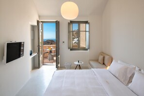 A modern bedroom with a large bed, a television, a small table, and a window with a view of a coastal landscape. - Old Castle Oia by Mr and Mrs White (Oia)