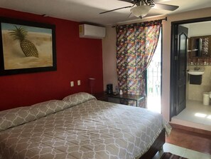 Quarto com cama, ventilador de teto, pintura de abacaxi, cortina colorida e banheiro visível por uma porta aberta.