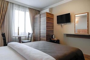 Quarto de hotel com uma cama grande, televisão fixada na parede, uma pequena mesa com cadeira e um guarda-roupa.