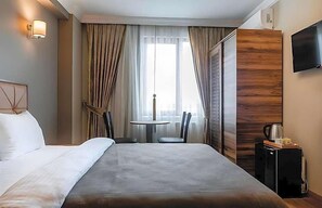 A hotel room with a bed, a small table with chairs, a TV mounted on the wall, and a window with curtains.