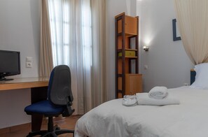 A hotel room with a bed, a desk with a computer, a chair, a wooden cabinet, and a window with curtains. - Porto Vecchio Luxury Suites (Spetses)