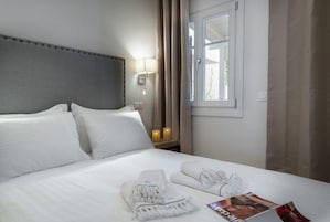 Una habitación de hotel con una cama, almohadas, una mesita de noche con una lámpara, una ventana con cortinas y una revista sobre la cama.
