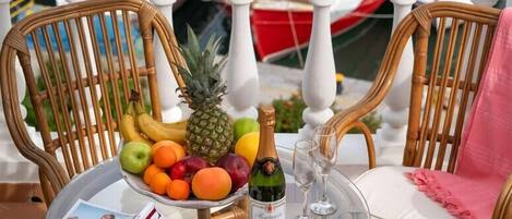 Mesa com uma cesta de frutas, uma garrafa de champanhe e uma revista sobre um tampo de vidro, cadeiras de vime e vista de barcos ao fundo.