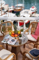 A table with a fruit bowl, a bottle of champagne, and a magazine on a glass table with wicker chairs and a view of boats in the background.