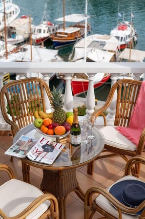 Una mesa redonda de vidrio con sillas de mimbre, un frutero, una botella de champán y una revista en una terraza con vistas a un puerto deportivo con barcos.
