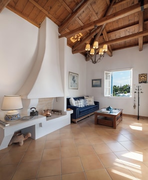 A spacious living room with a high wooden ceiling, a white fireplace, a blue sofa, and a large window offering a view of greenery.