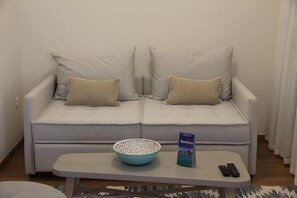 A white sofa with two beige pillows, a blue bowl, a magazine, and two remote controls on a small table. - Porto Vecchio Luxury Suites (Spetses)