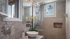 Un baño con pared de piedra, un lavamanos blanco, un espejo, una ducha y un estante pequeño con artículos de tocador.