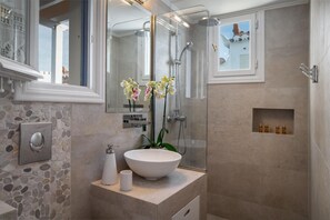 A bathroom with a stone wall, a white sink, a mirror, a shower, and a small shelf with toiletries. - Porto Vecchio Luxury Suites (Spetses)