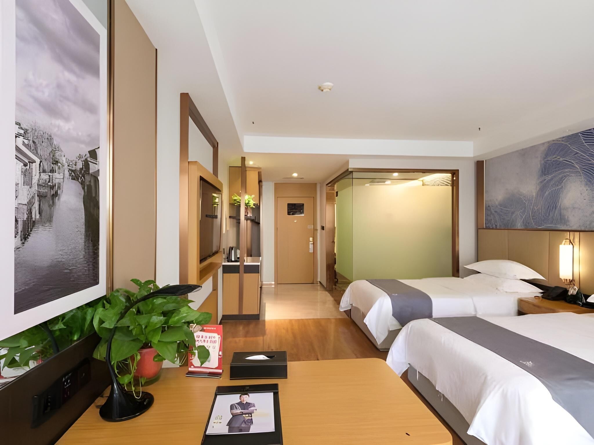 A hotel room with two beds, a desk with a computer, a potted plant, and artwork on the walls.