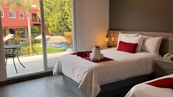 Una habitación de hotel moderna con dos camas, un balcón y vistas a un edificio rojo y vegetación.