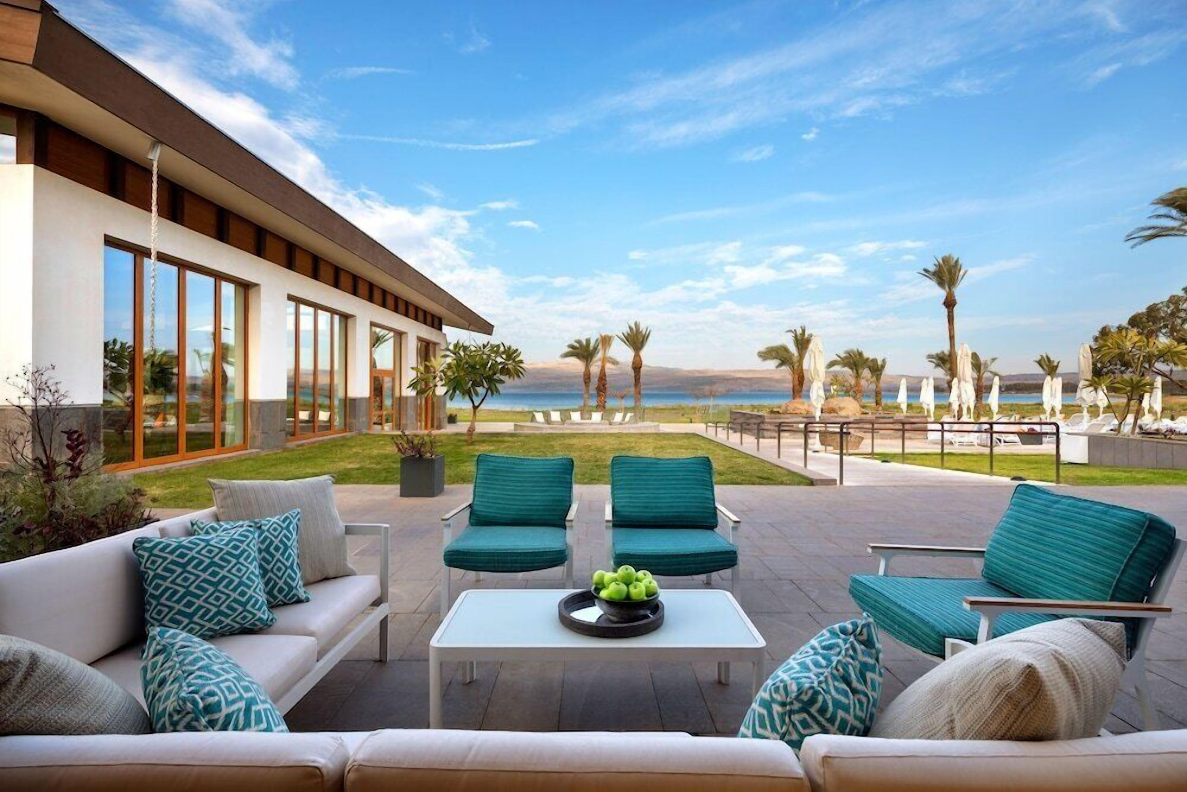 A modern outdoor seating area with a view of the sea, featuring a white sofa, two green armchairs, and a coffee table with a fruit bowl.