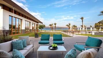 A modern outdoor seating area with a view of the sea, featuring a white sofa, two green armchairs, and a coffee table with a fruit bowl.