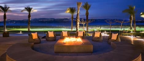 A circular outdoor seating area with a fire feature, surrounded by palm trees and overlooking a body of water.