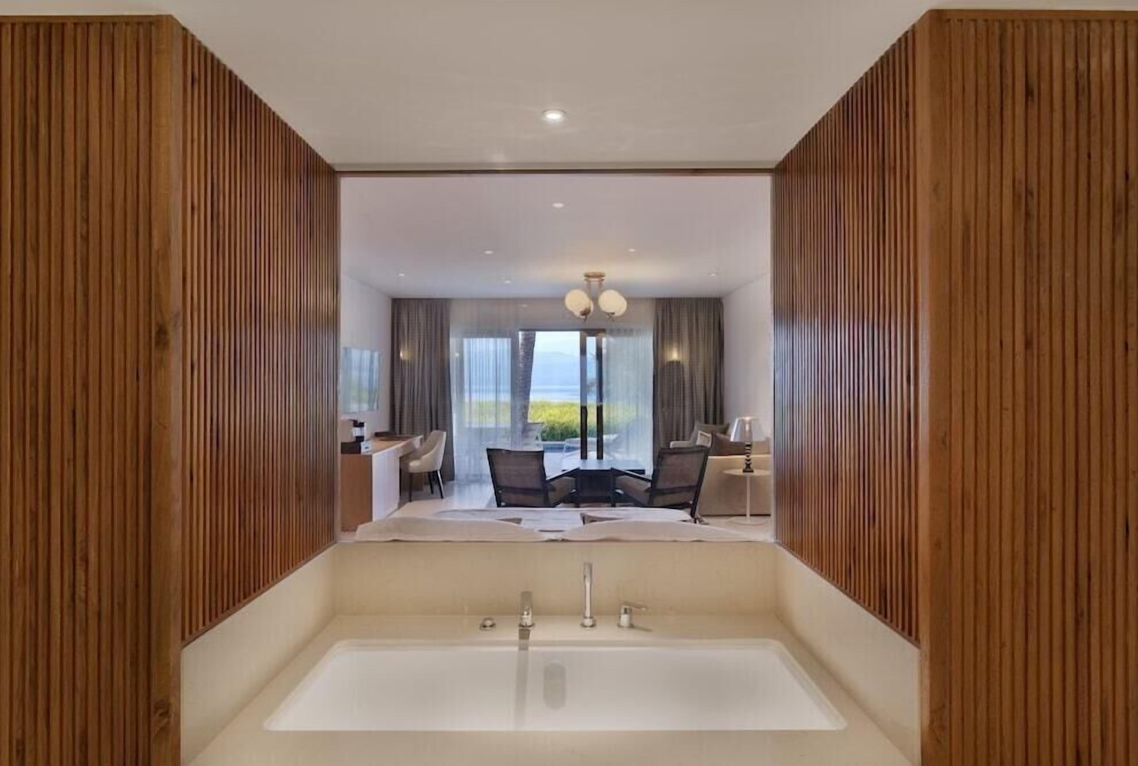 A modern bathroom with a large bathtub, wooden paneling, and a view of a dining area with a table and chairs.