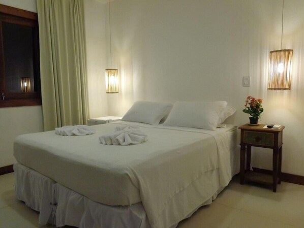 Una cama bien hecha con sábanas y toallas blancas, una mesita de noche con un jarrón de flores y dos lámparas fijadas a la pared.