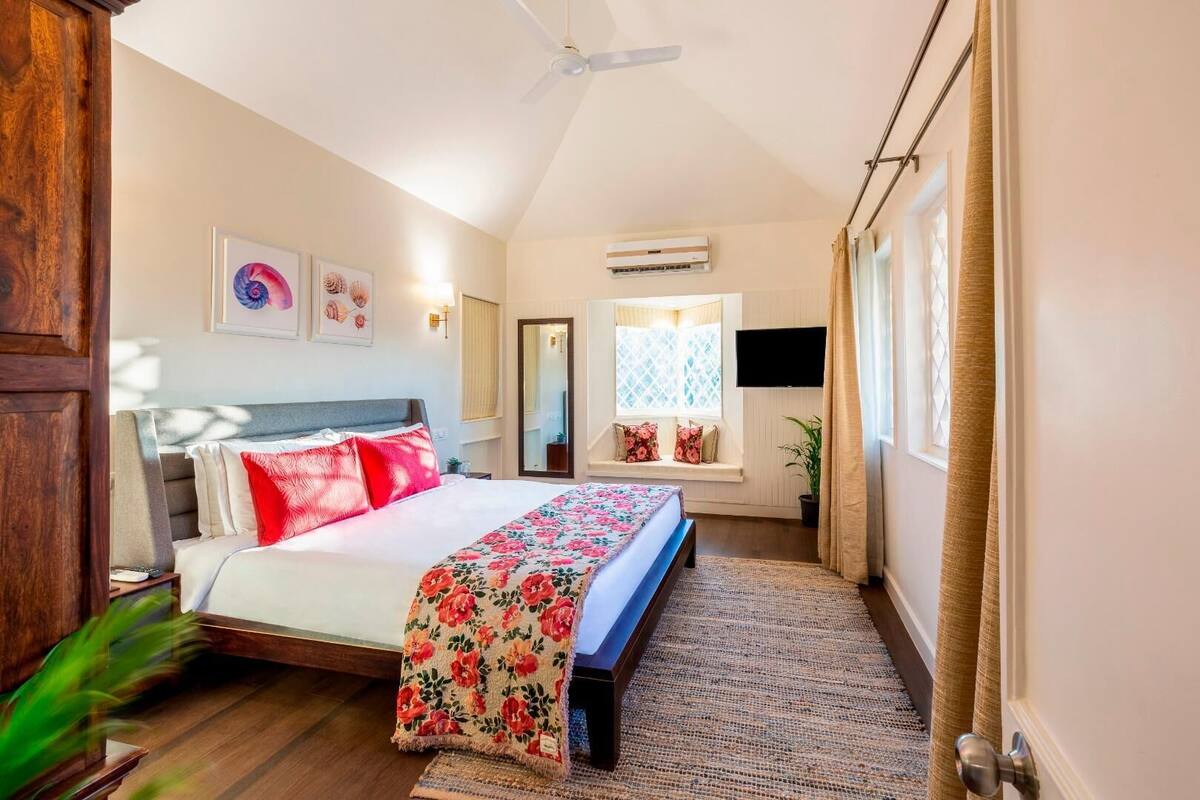 A bedroom with a bed, a ceiling fan, an air conditioning unit, a window with curtains, and a wall-mounted TV.