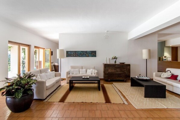 Una sala de estar moderna con dos sofás, una mesita de centro y un aparador. Hay ventanas grandes y un cuadro en la pared.
