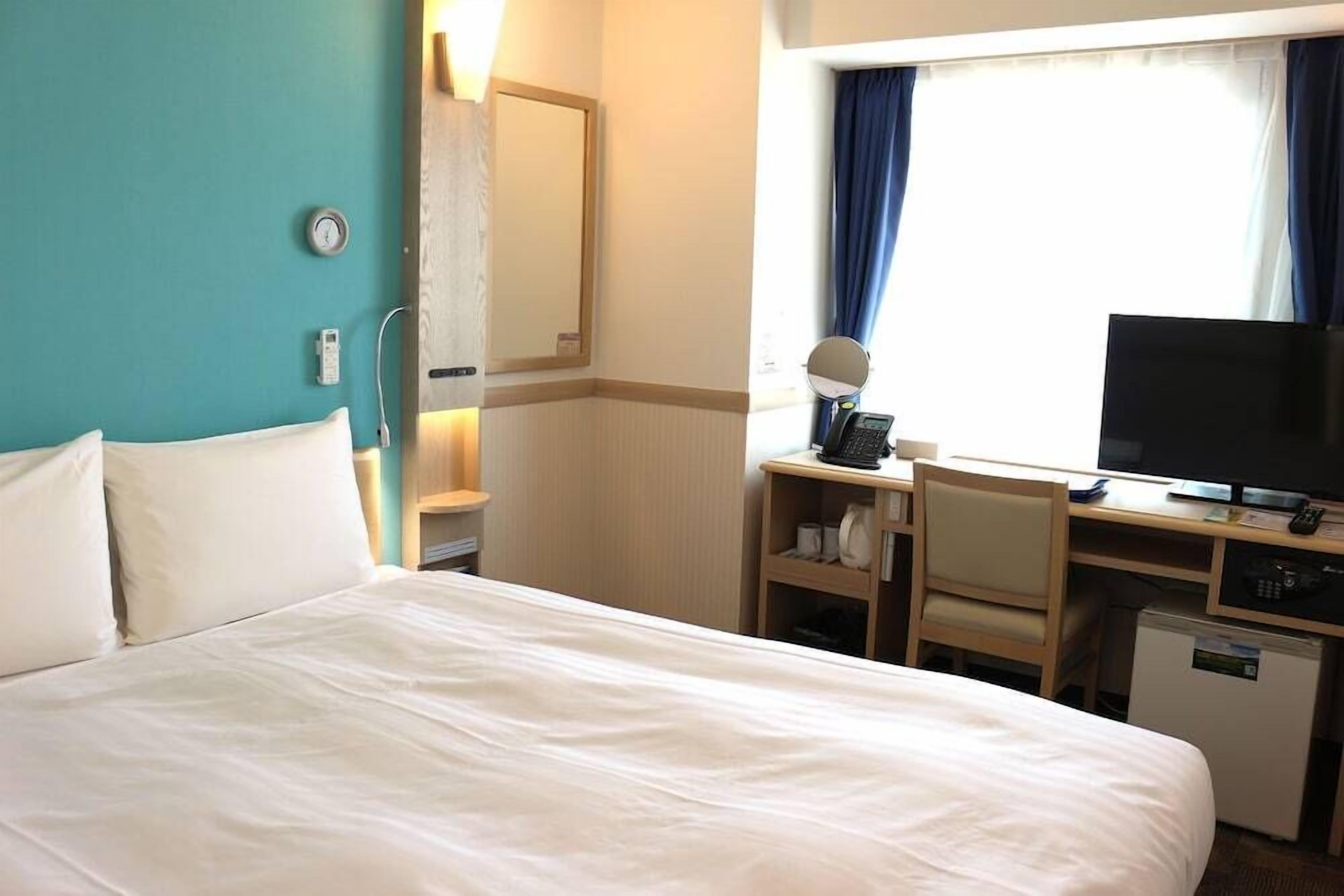 A hotel room with a bed, a desk with a computer, a television, and a small refrigerator.