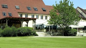 A large building with a red-tiled roof, multiple windows, and a covered outdoor area with chairs and tables.