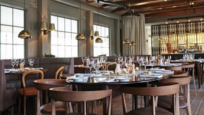 A restaurant with round tables set for dining, large windows, and hanging pendant lights.