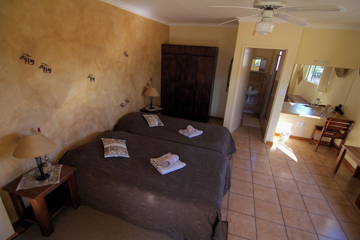 A hotel room with two beds, a wooden wardrobe, a bathroom visible through an open door, and a small kitchenette area.