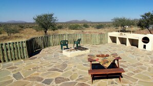 An outdoor patio with stone flooring, two green chairs, a wooden table with a patterned tablecloth, and a stone fire pit. - Etango Ranch Guest Farm (Windhoek)