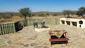 An outdoor patio with stone flooring, two green chairs, a wooden table with a patterned tablecloth, and a stone fire pit. - Etango Ranch Guest Farm (Windhoek)